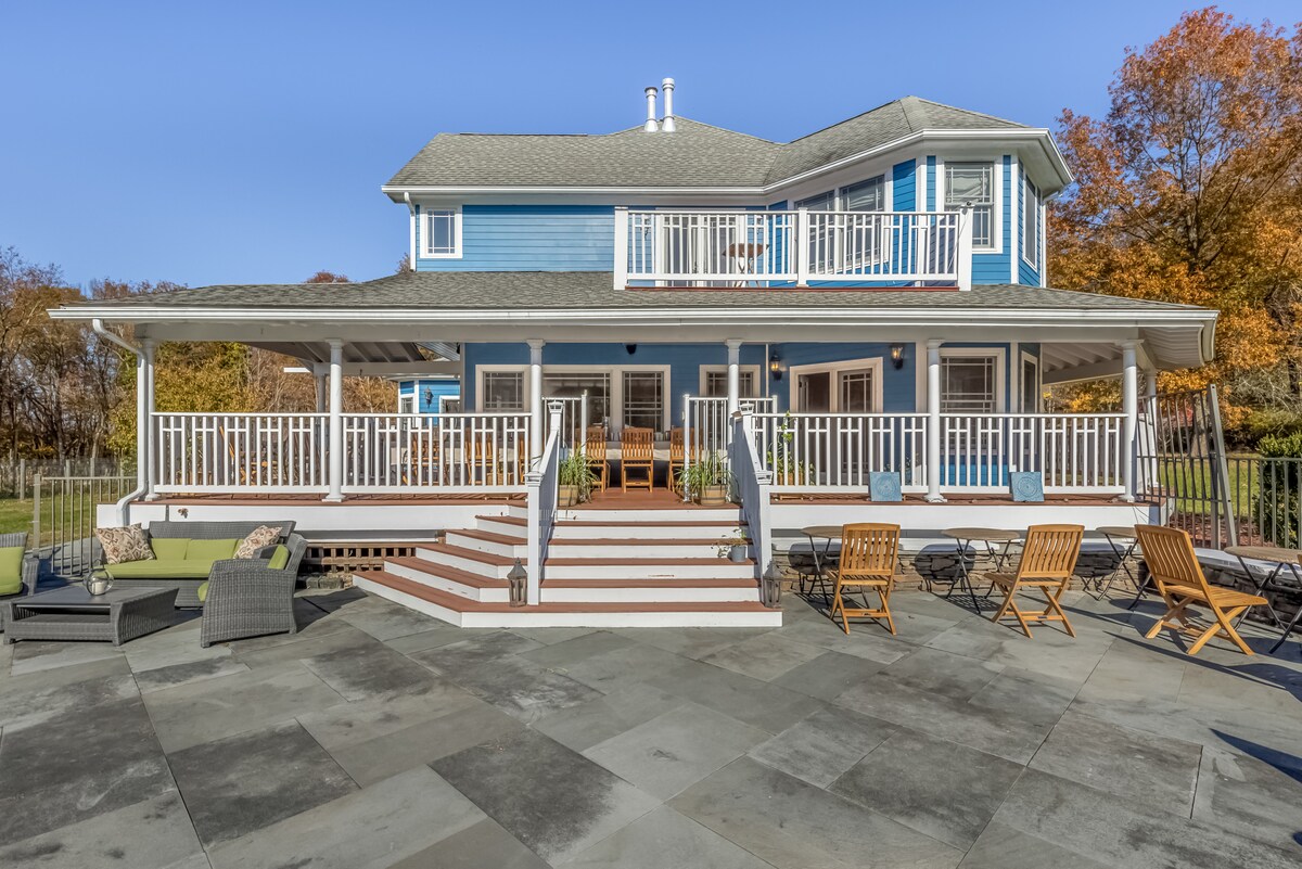Thee Wild Lux estate facade from the pool terrace — blue house, wrap-around porch, outdoor seating