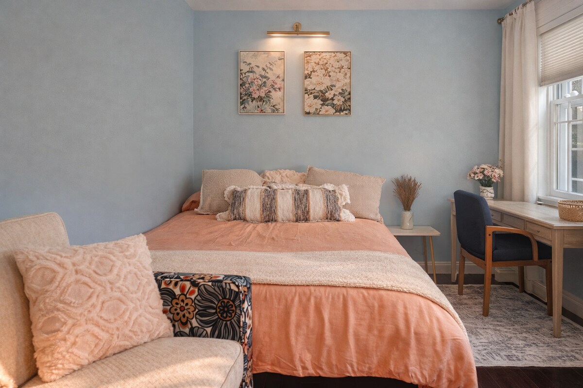 The Blue Room at Thee Wild Lux — soft blue walls, queen bed with warm linen, floral botanical art above headboard